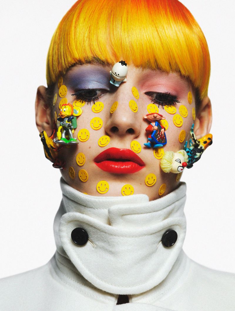 Close-up portrait of a person with yellow hair and smiley face stickers on their face, wearing a white coat with black buttons.
