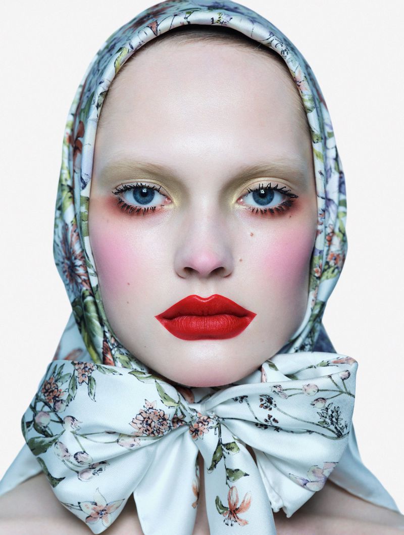 Close-up portrait of a fair-skinned woman with bright red lipstick and heavy blush, wearing a floral-patterned headscarf, on a white background.