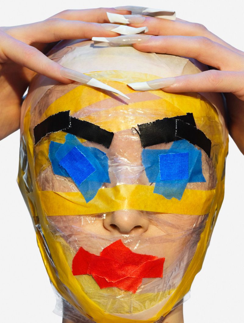 Person with artful tape covering face, hands posed near head. Close-up on white background. Creative beauty portrait, bold color, expressive.