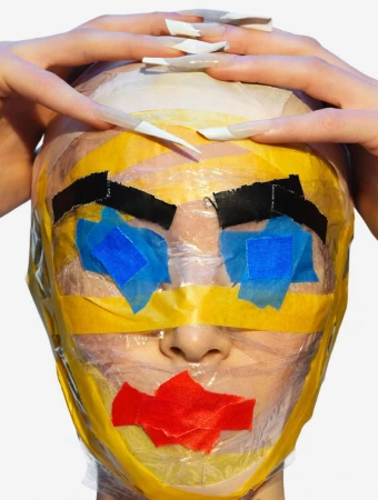 Person with artful tape covering face, hands posed near head. Close-up on white background. Creative beauty portrait, bold color, expressive.