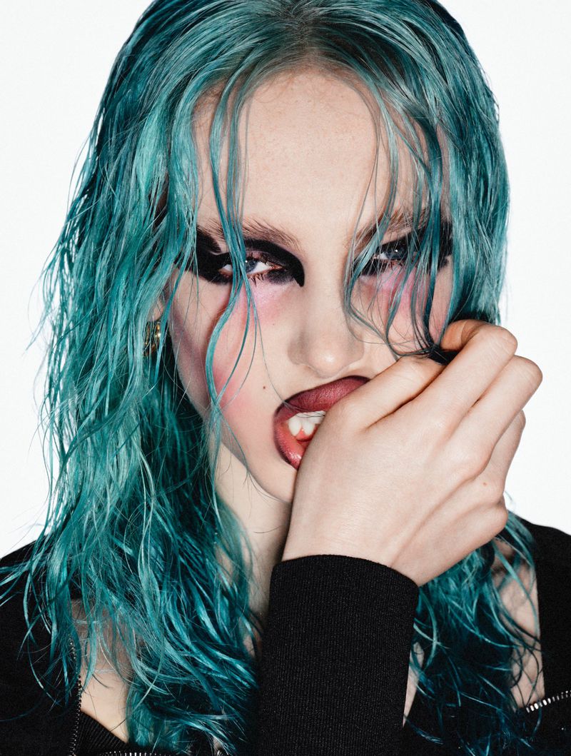 Close-up portrait of a young adult woman with wet blue hair, dark eye makeup, and an open mouth, against a white background.
