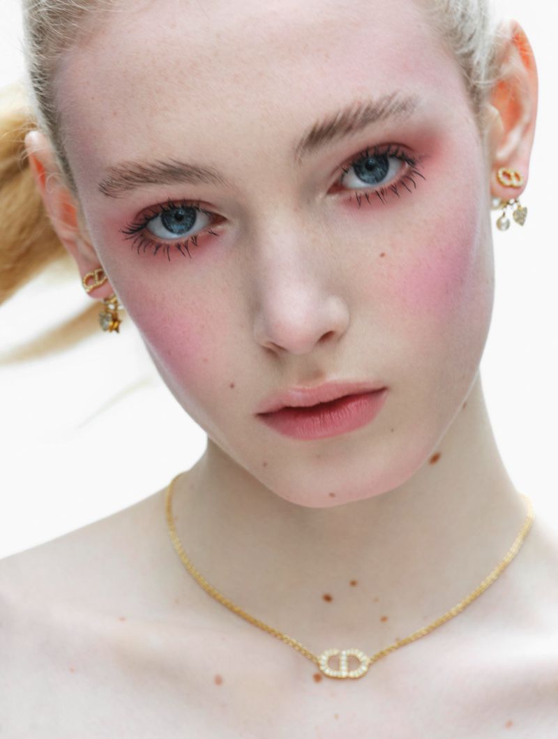 Close-up studio shot of a young, fair-skinned woman modeling earrings and a necklace with the CD logo, wearing pink blush.
