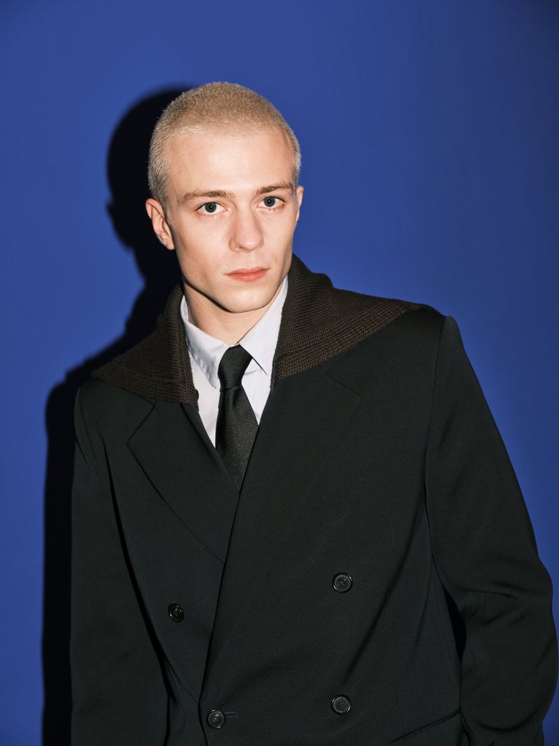 Young man in a black double-breasted suit and black tie, looking at the camera against a blue background with a shadow.