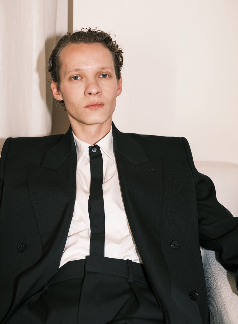 Portrait of a fair-skinned adult male in a black suit and white shirt, sitting indoors with a neutral expression and natural lighting.