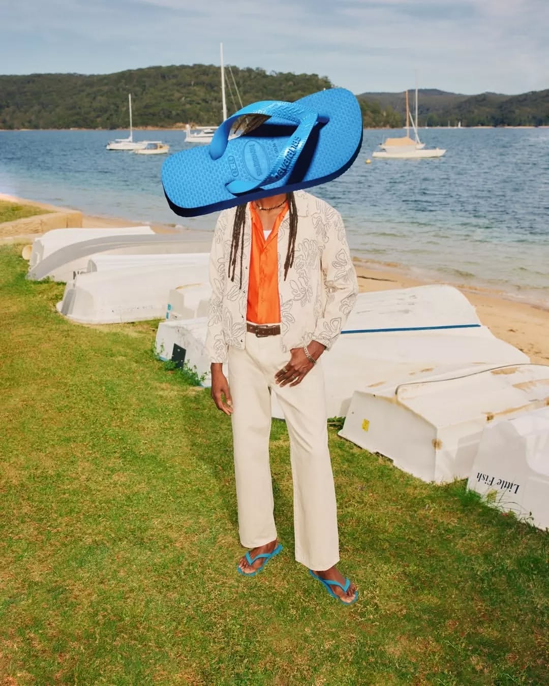 Man stands on grassy beach with sailboats; he is wearing blue flip-flops on his feet and balanced on his head as a hat.