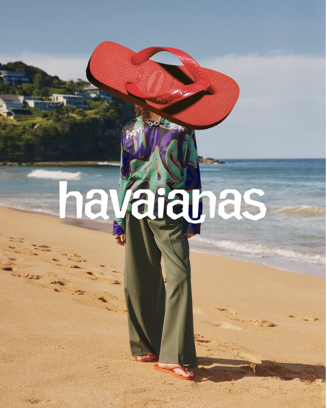 Person in trendy green pants and patterned top standing on a beach under a large, superimposed red Havaianas flip-flop and logo.