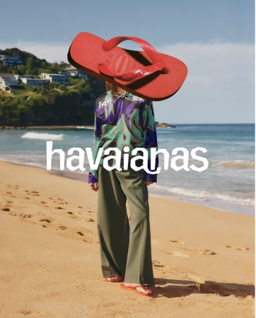 Person in trendy green pants and patterned top standing on a beach under a large, superimposed red Havaianas flip-flop and logo.