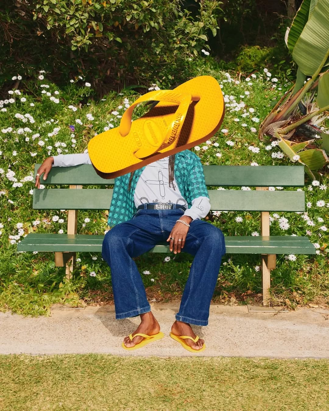 Person sits on park bench with yellow flip-flops obscuring face, wearing jeans, patterned shirt and a white t-shirt in outdoor setting.