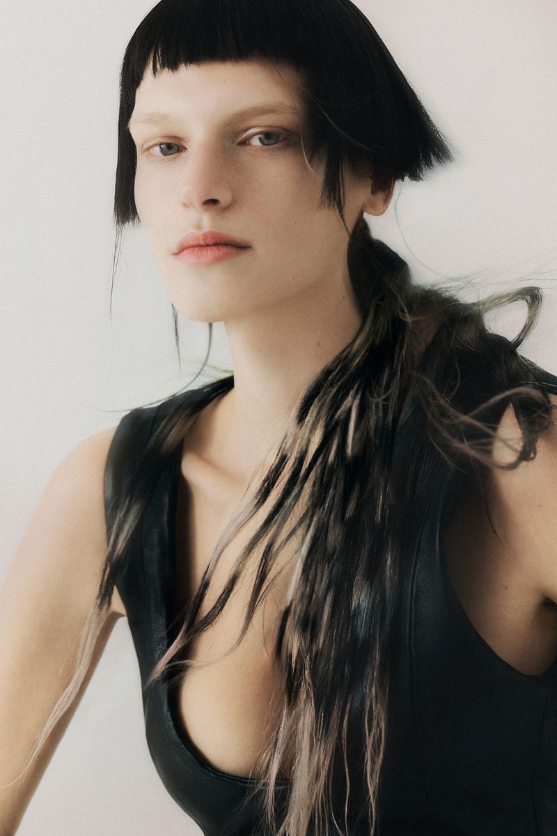 A fair-skinned adult female model with a blunt bob and low ponytail poses in a sleeveless black top against a neutral background.