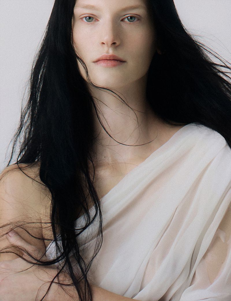 Portrait of an adult female with long, dark hair and pale skin wearing a white garment, set against a light neutral background.