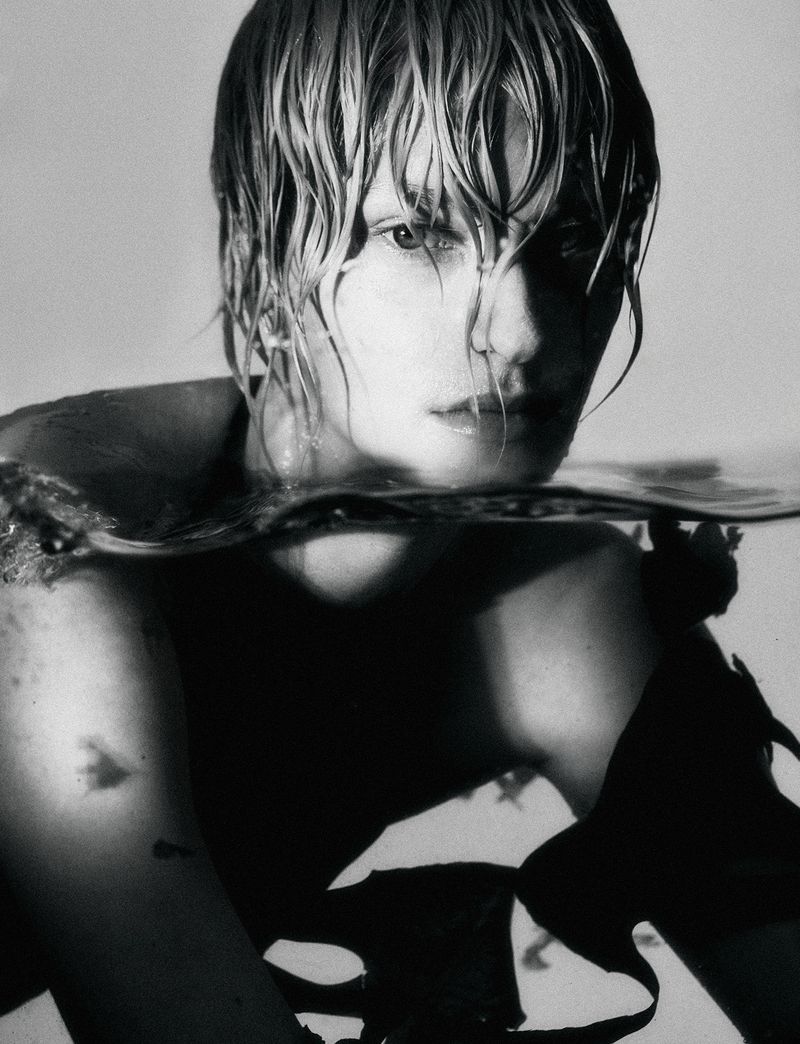 Monochrome portrait of a person partially submerged in water, showcasing wet hair, shoulders, and a serene facial expression against a light background.