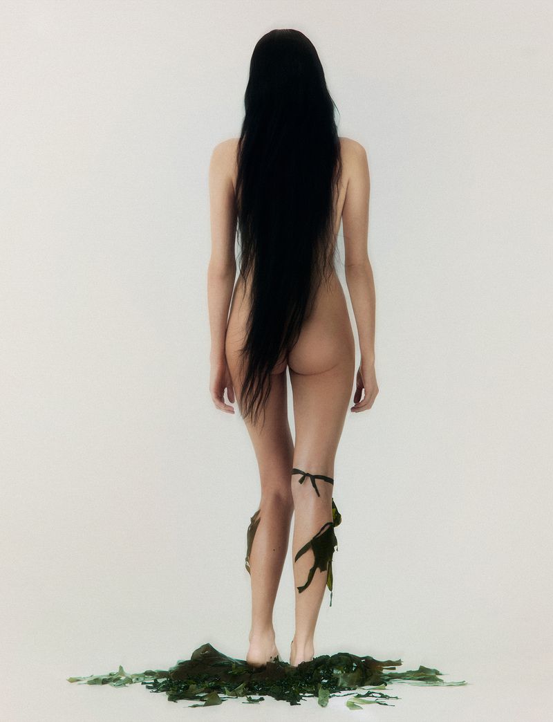A woman stands with long, dark hair, showcasing minimal adornment and organic textures against a plain backdrop, emphasizing natural aesthetics.