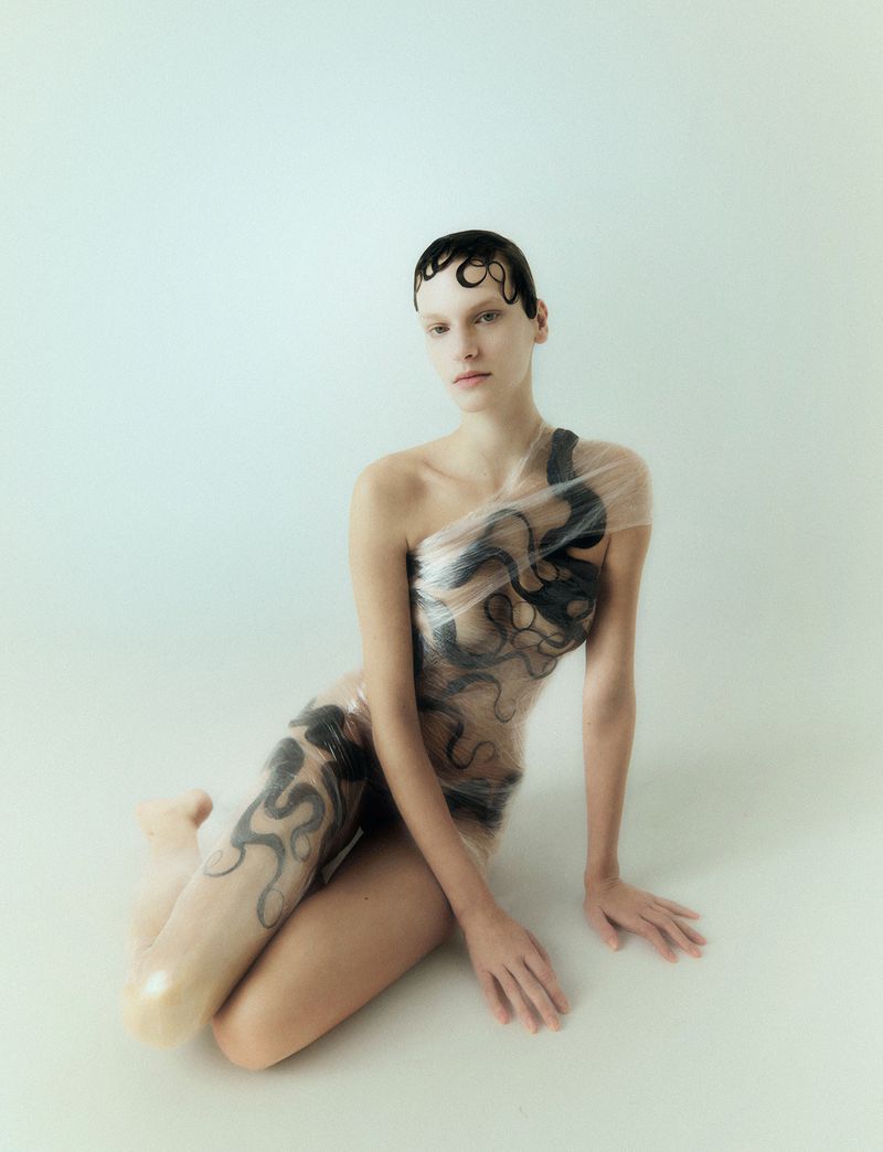 Artistic image of a woman with wet hair, sitting and wrapped in patterned plastic against a light background, showcasing a unique beauty style.