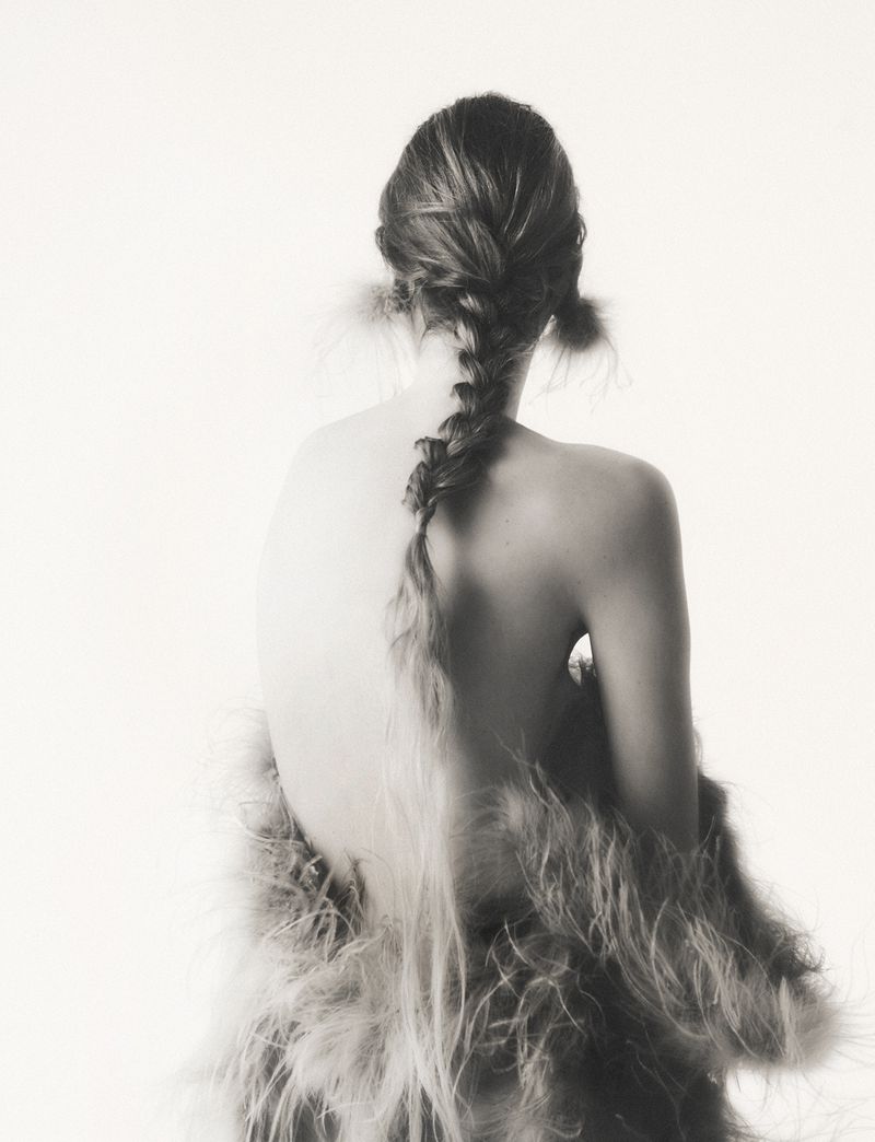 Back view of a woman with a long braid wearing a feathered shawl against a white background in a high-key, monochromatic photograph.
