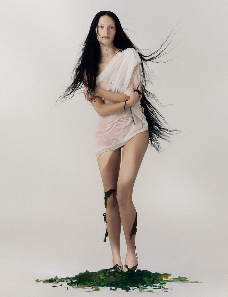 A woman with long dark hair stands in a sheer white dress surrounded by green seaweed in a studio setting against a white background.