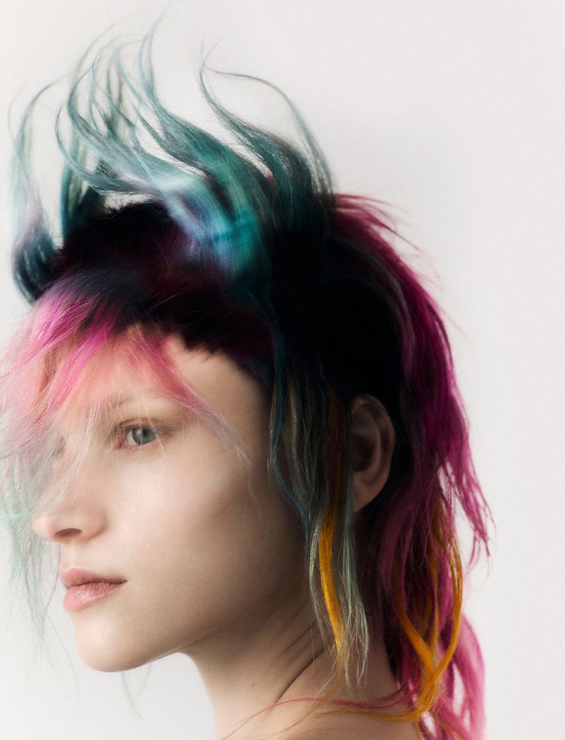 Close-up portrait of a woman with vibrant, multicolored hair in teal, pink, and yellow against a light backdrop in a beauty-style photograph.