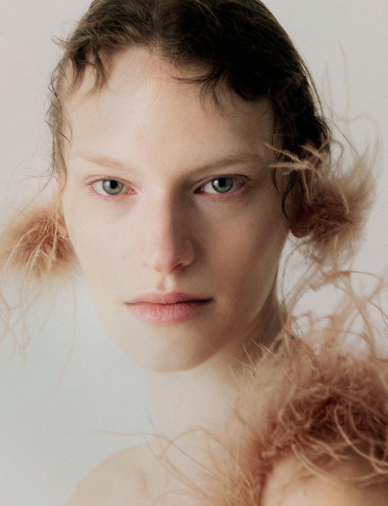 Close-up portrait of a woman with fair skin and light hair, adorned with feathers around her face, set against a bright background.