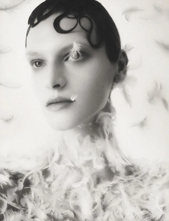 Monochromatic portrait of a fair-skinned young adult woman with dark, stylized hair, adorned with white feathers, against a white backdrop.