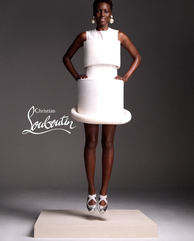 Fashion shot of a Black woman in a white dress, jumping on a white platform in a grey photo studio with 'Christian Louboutin' logo