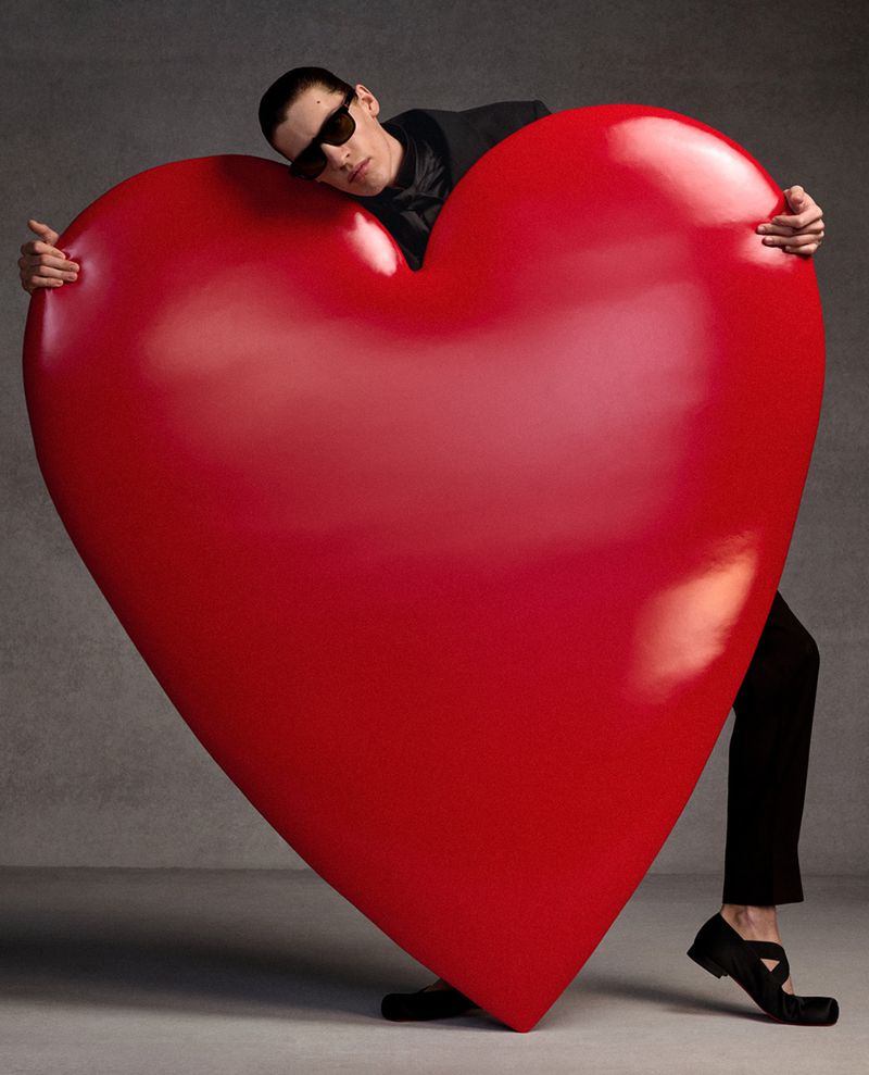 Fashionable adult male with sunglasses leans against a large red heart. Man is wearing a black suit and black shoes in a studio.