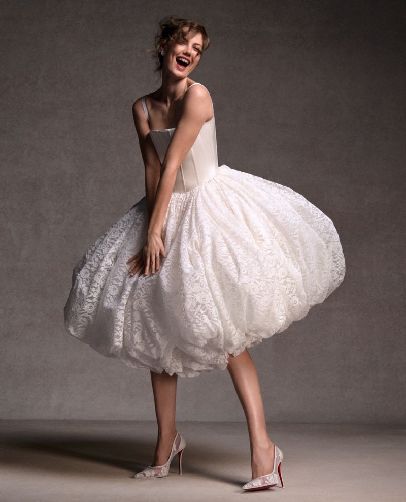 Smiling woman in a white dress with a voluminous skirt and white and red high heel shoes in a studio with a grey backdrop.