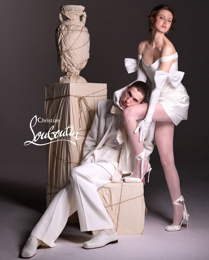 Fashion shot of a couple in all-white attire posing in a studio setting. Brand logo visible. Sculpture and art installation included.