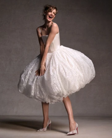 Smiling white woman wearing a white lace bubble dress and high heels against a grey backdrop