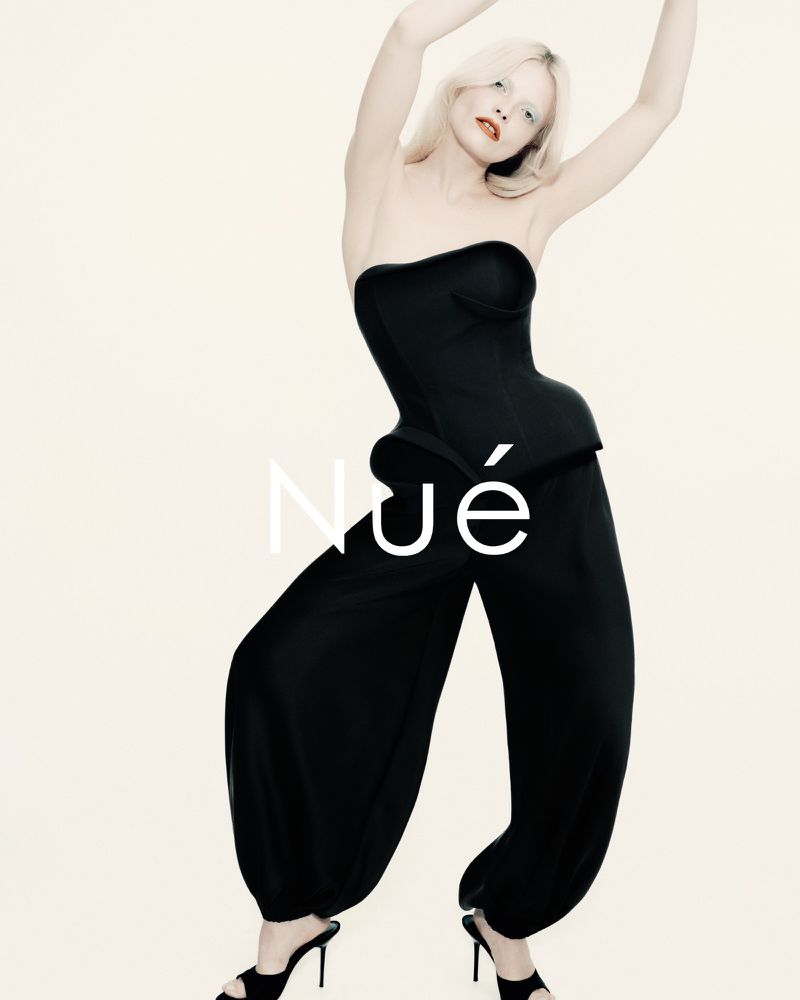 A single, blonde female model wearing a black strapless corset top and harem pants, poses with arms raised in a light studio.