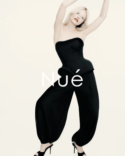 A single, blonde female model wearing a black strapless corset top and harem pants, poses with arms raised in a light studio.
