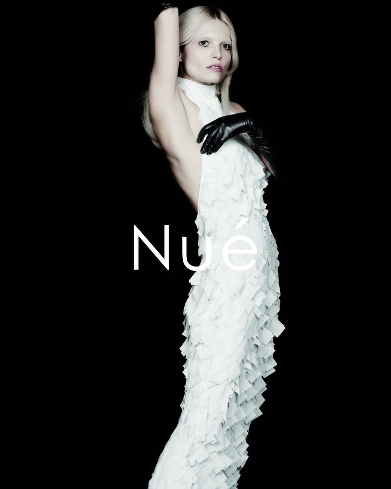 Fashion portrait of a woman with platinum blonde hair wearing a white dress with cascading ruffles and black leather gloves against black backdrop.