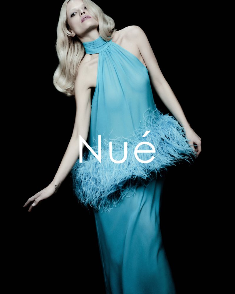 Model in a teal halter dress with feather trim posing against a black background for NUÉ Resort 2026