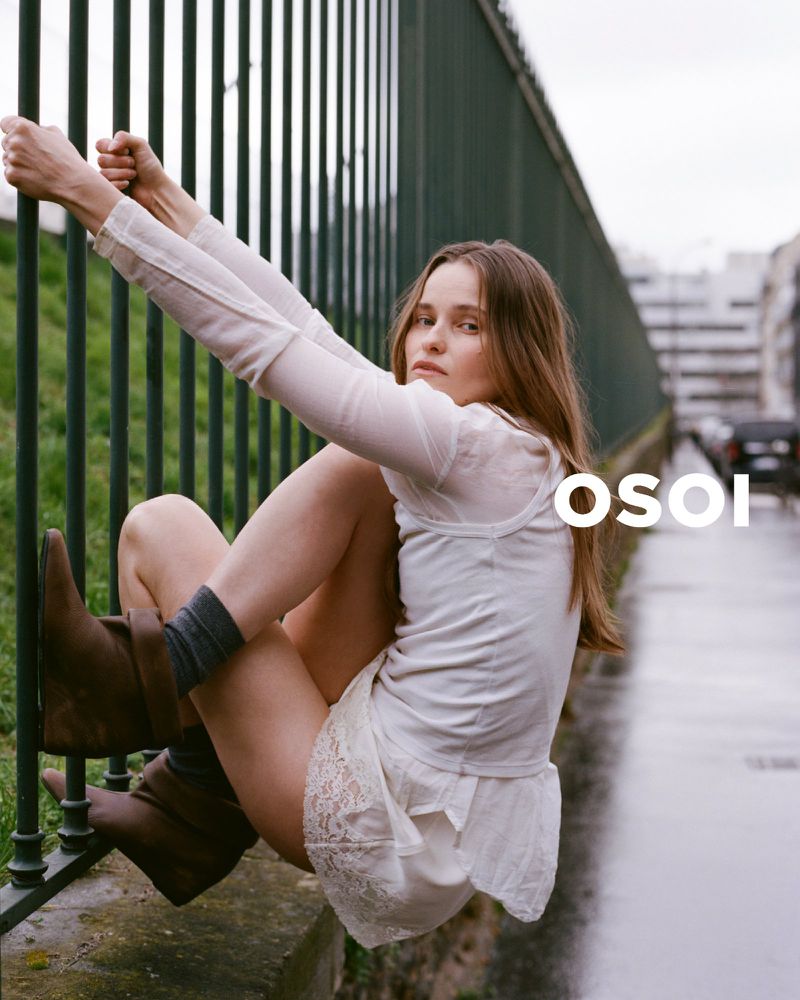A young woman with long hair sits on a green fence wearing a white top and skirt with brown boots in an outdoor setting.