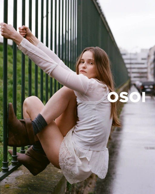 A young woman with long hair sits on a green fence wearing a white top and skirt with brown boots in an outdoor setting.