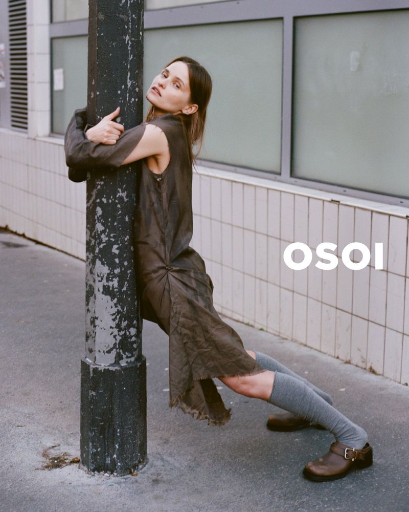 A woman in a draped dress hugs a utility pole on a city street. She wears knee-high socks and clogs. White graphic text.