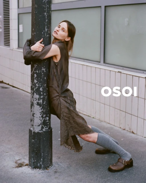 A woman in a draped dress hugs a utility pole on a city street. She wears knee-high socks and clogs. White graphic text.