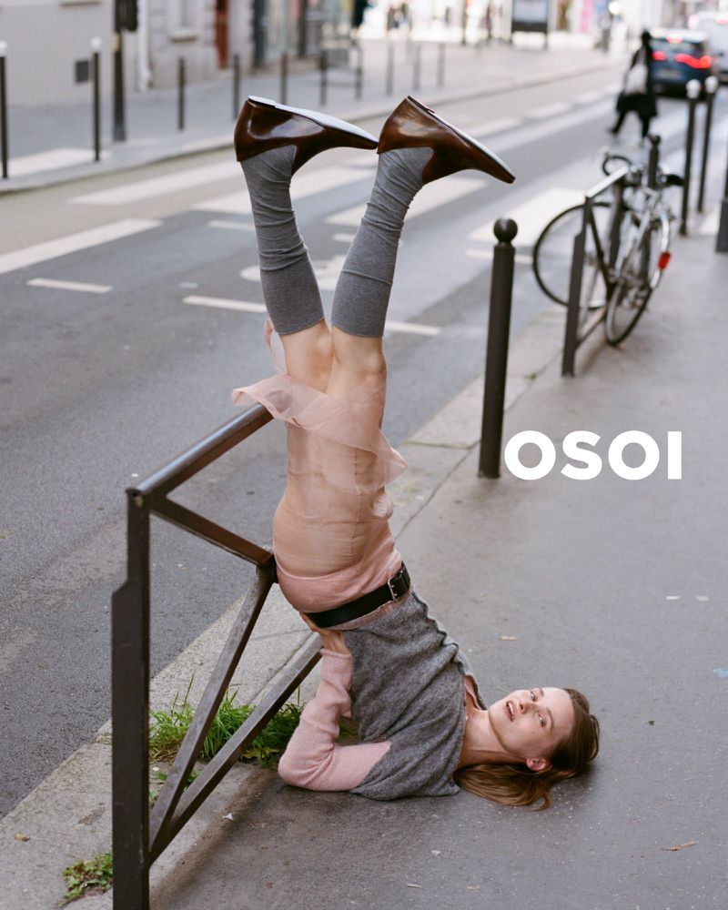 Fashion shot of a woman upside down, balancing on a handrail wearing brown shoes, grey socks, and a pink skirt on a city sidewalk.