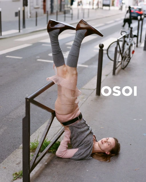 Fashion shot of a woman upside down, balancing on a handrail wearing brown shoes, grey socks, and a pink skirt on a city sidewalk.