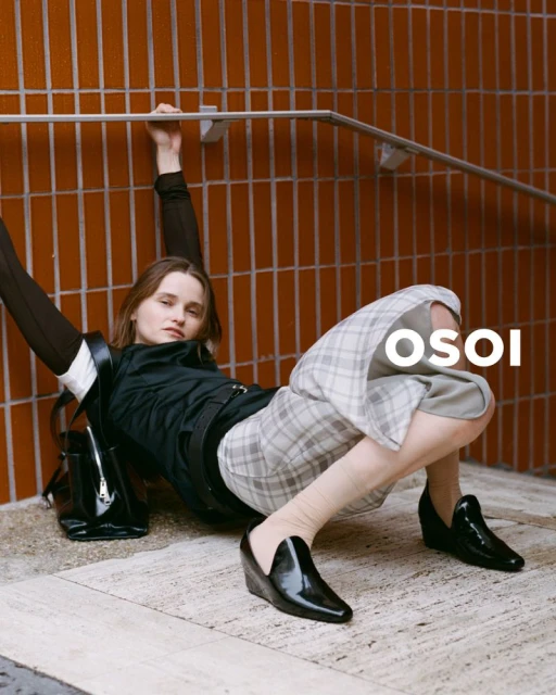 Woman in black top, plaid shorts, and patent shoes leans back against an orange tiled wall with hand on a silver railing.