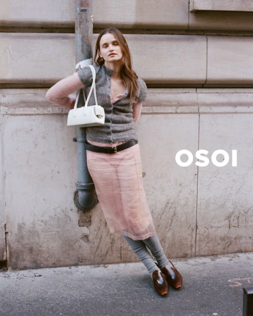 Woman in sweater and skirt stands next to a weathered wall, holding a handbag, in an outdoor fashion shot with natural lighting.