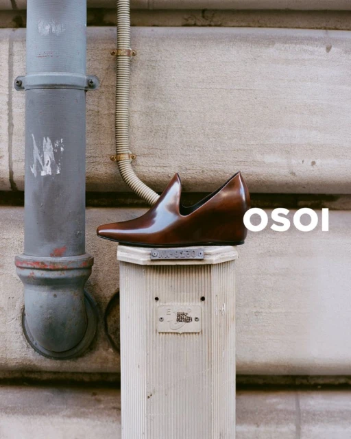 Brown leather OSOI shoe sitting on a short, ridged white pillar with gray pipes and neutral wall in the background; studio product shot.