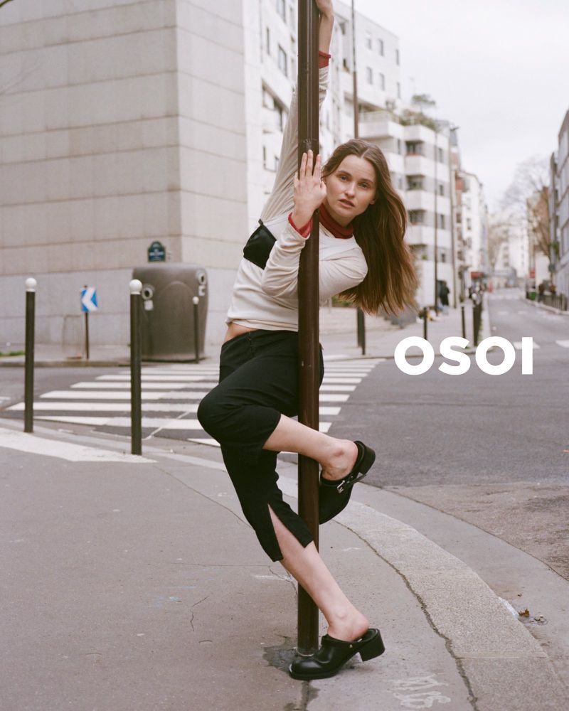 Fashion photo of a young woman leaning against a pole on a city street, wearing a long-sleeved top, black pants, and black clogs.