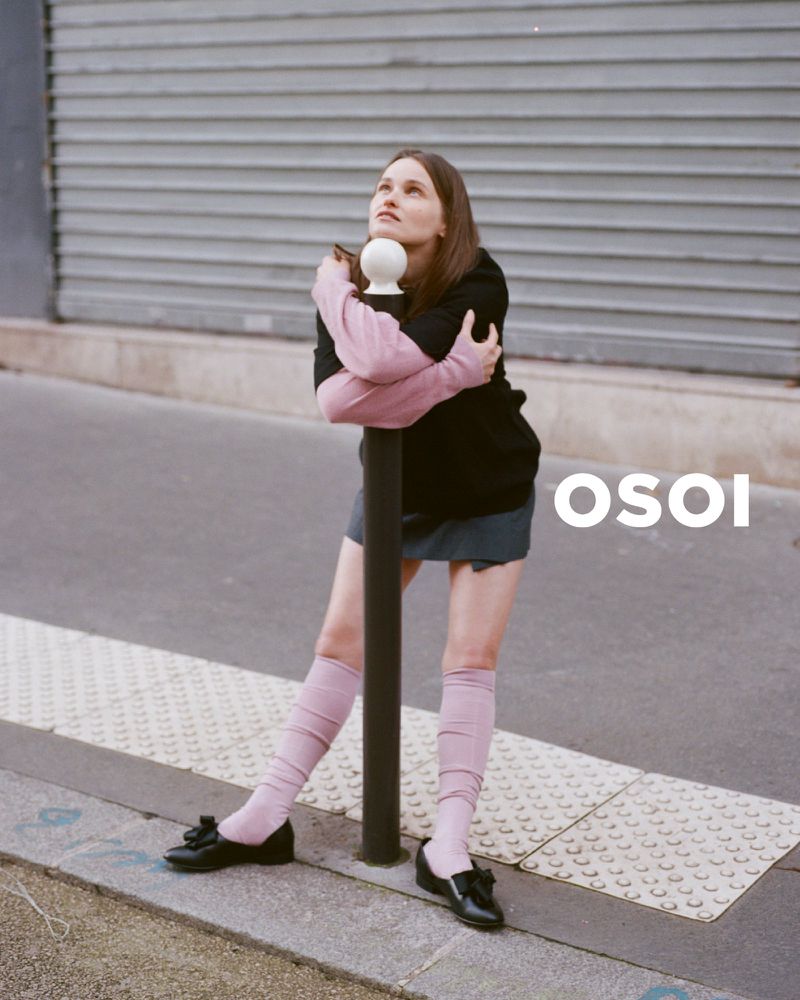 A woman hugging a black bollard on a city street wearing a black top, skirt, pink socks, and black shoes.