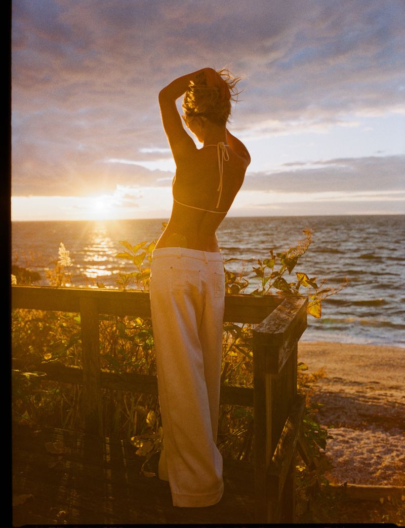 Woman in a bikini and white pants, standing on wooden steps at a beach with a sunset and ocean view in the background.