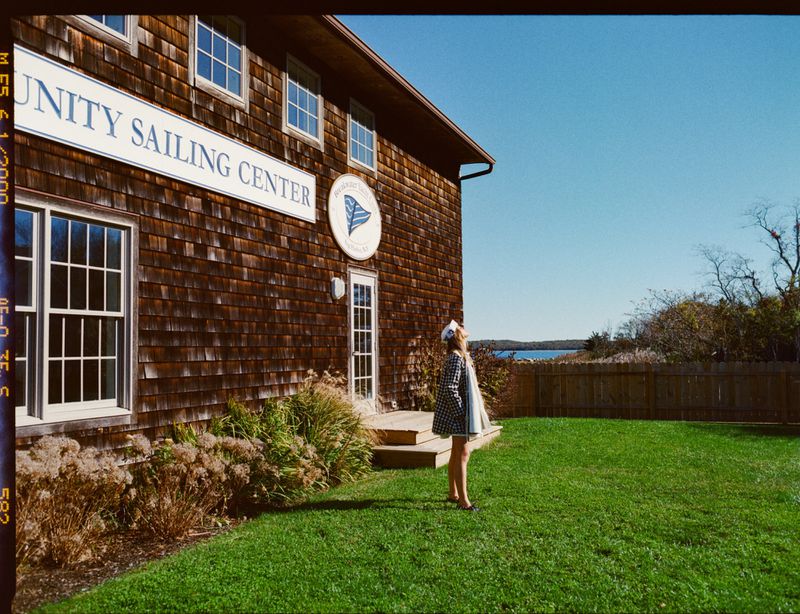 Young woman stands on green lawn in front of the Unity Sailing Center with its cedar-shingled facade on a sunny day near the water.