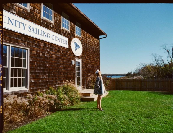 Young woman stands on green lawn in front of the Unity Sailing Center with its cedar-shingled facade on a sunny day near the water.