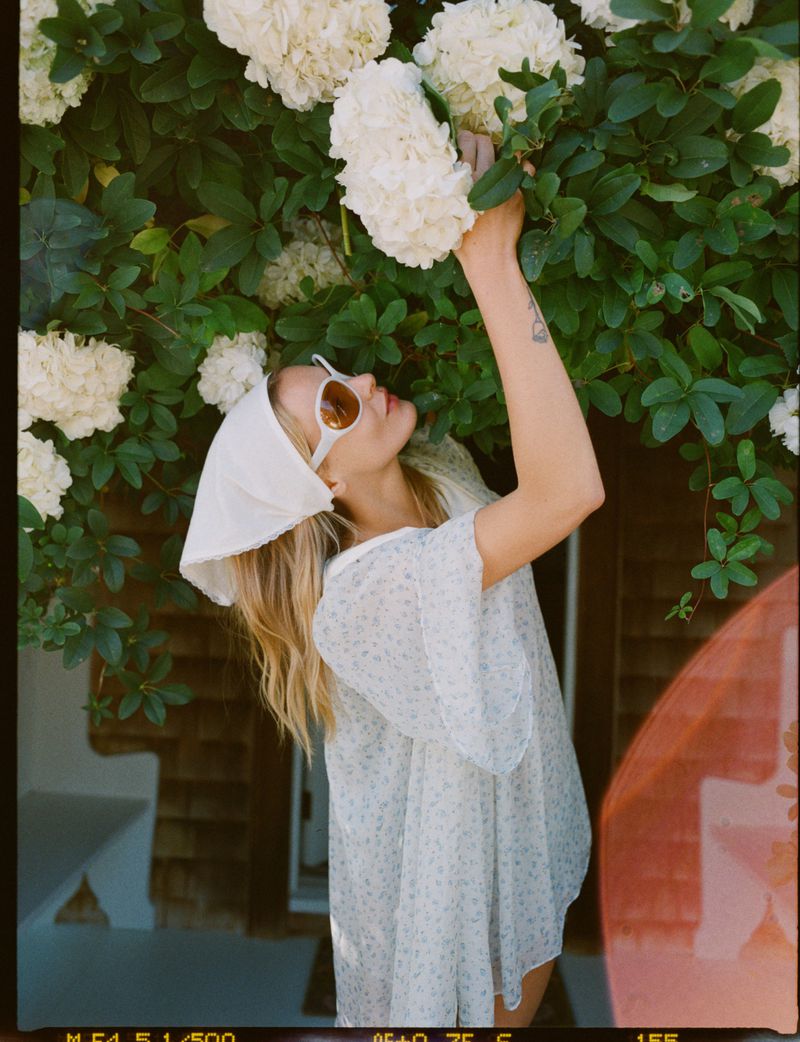 Woman in white headscarf and sunglasses reaches for white flowers on a leafy bush in natural light with a residential building in the background.