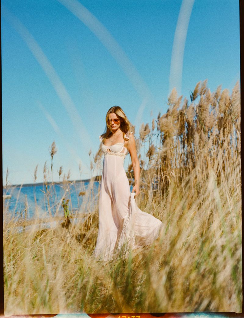 Woman in a dress walking through tall grass near the ocean on a sunny day. She has sunglasses and long wavy hair.