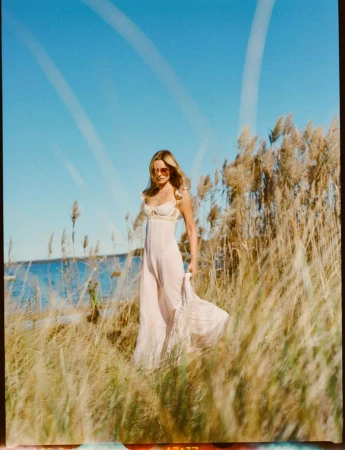 Woman in a dress walking through tall grass near the ocean on a sunny day. She has sunglasses and long wavy hair.