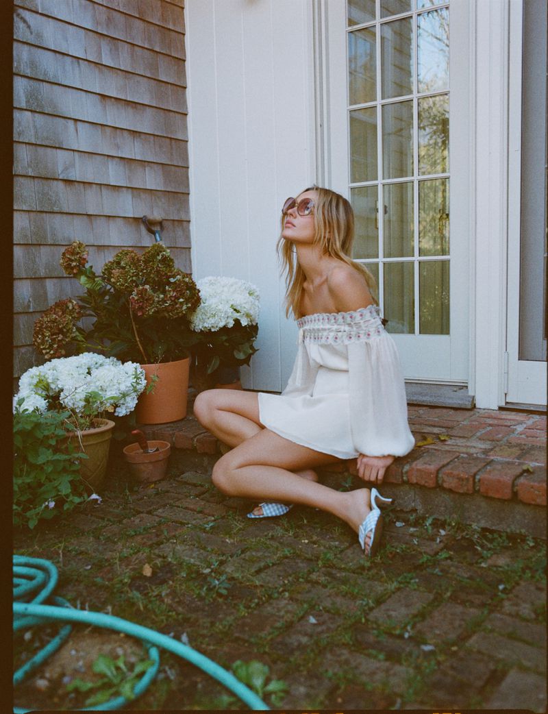 Blonde woman in white off-shoulder dress sitting on brick patio near white hydrangeas and green hose, looking up wearing pink sunglasses.