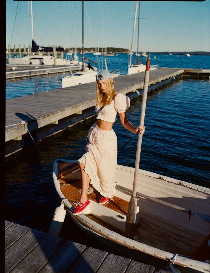 A blonde woman in a matching set stands on a small boat at a dock on a sunny day, posing with an oar and wearing a baseball cap.